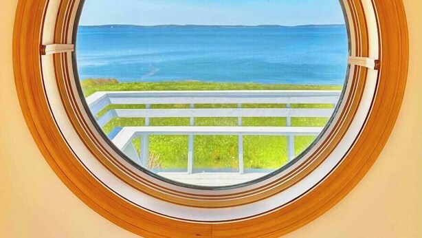 Una ventana redonda con marco de madera, con vistas a un área cubierta de césped y una masa de agua.
