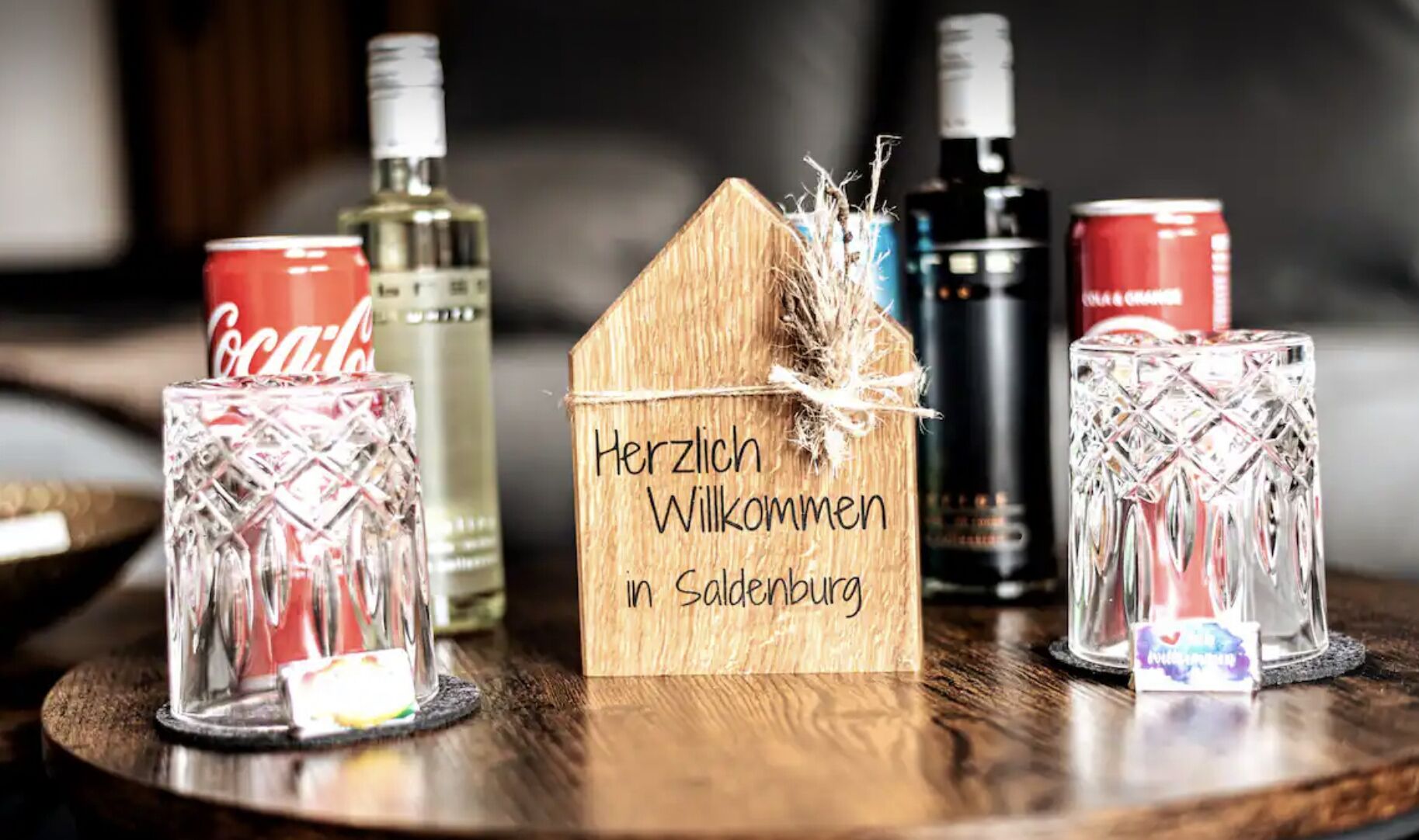 A wooden house-shaped sign with "Herzlich Willkommen in Salzburg" written on it, surrounded by various bottles and glasses on a wooden table.