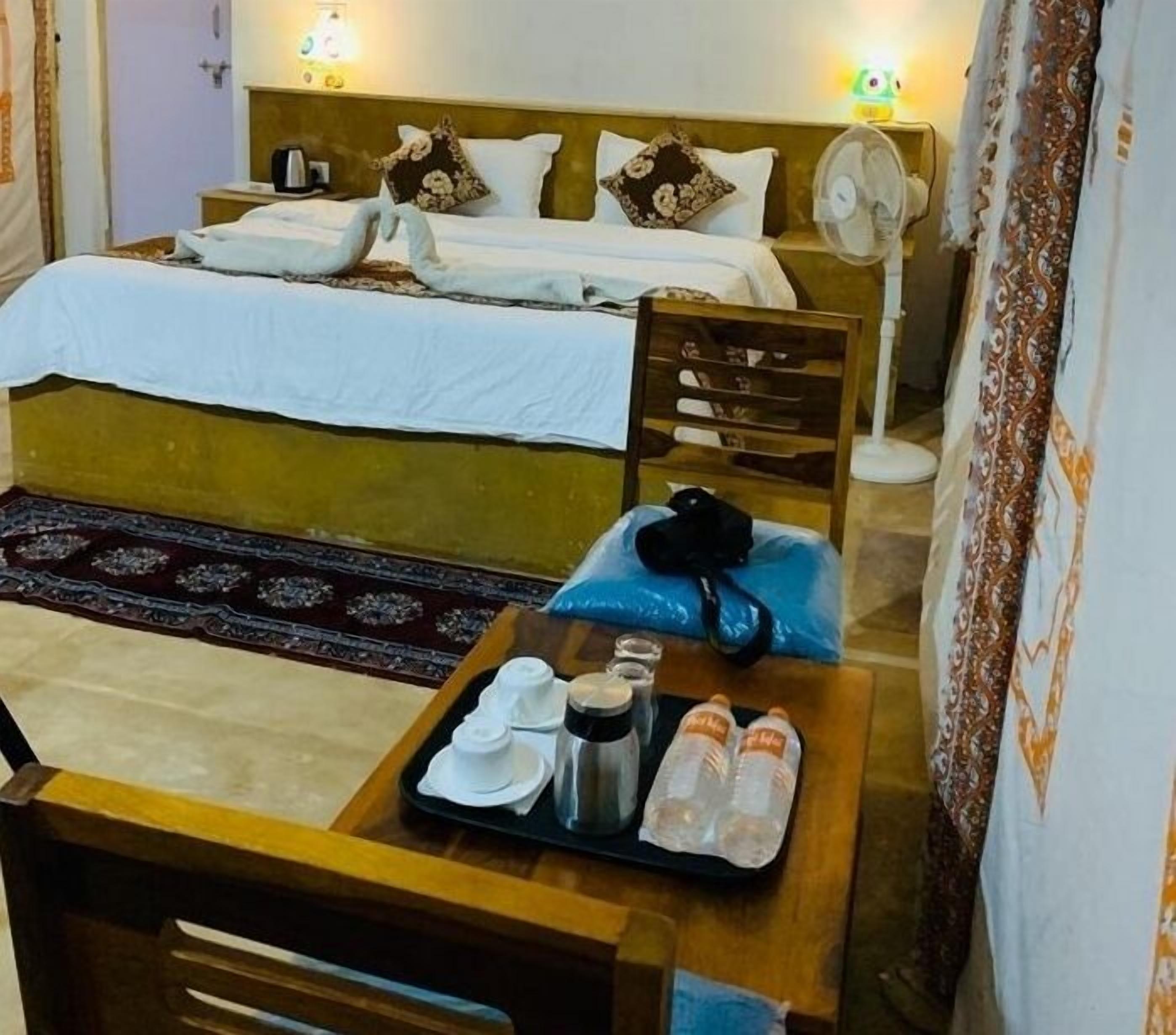 Una habitación de hotel con una cama, una silla de madera, una mesita con artículos de aseo y un ventilador.
