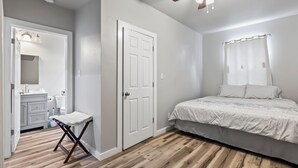 Une chambre avec un plancher en bois, un lit avec une literie blanche, une porte et une salle de bain équipée d’un lavabo et d’un miroir.