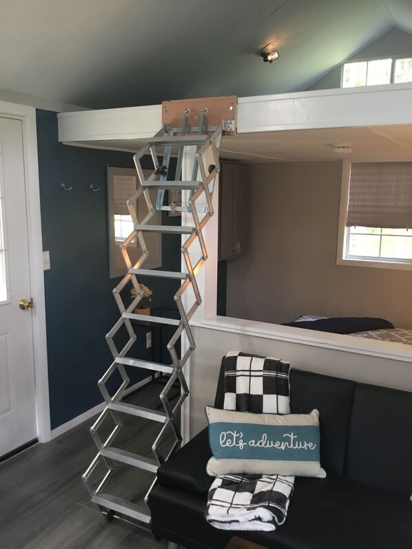 A room with a bunk bed, a ladder, a sofa with decorative pillows, a window, and a door.