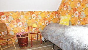 Et soveværelse med blomstret tapet, en seng, en stol, et lille bord med en lampe og en gul pude med tekst.