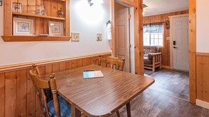 A wooden dining table with chairs, a wall-mounted shelf with glassware, and a door leading to another room. - Bluegill Bungalow-9 Miles To Dells, Pet Friendly (Briggsville)