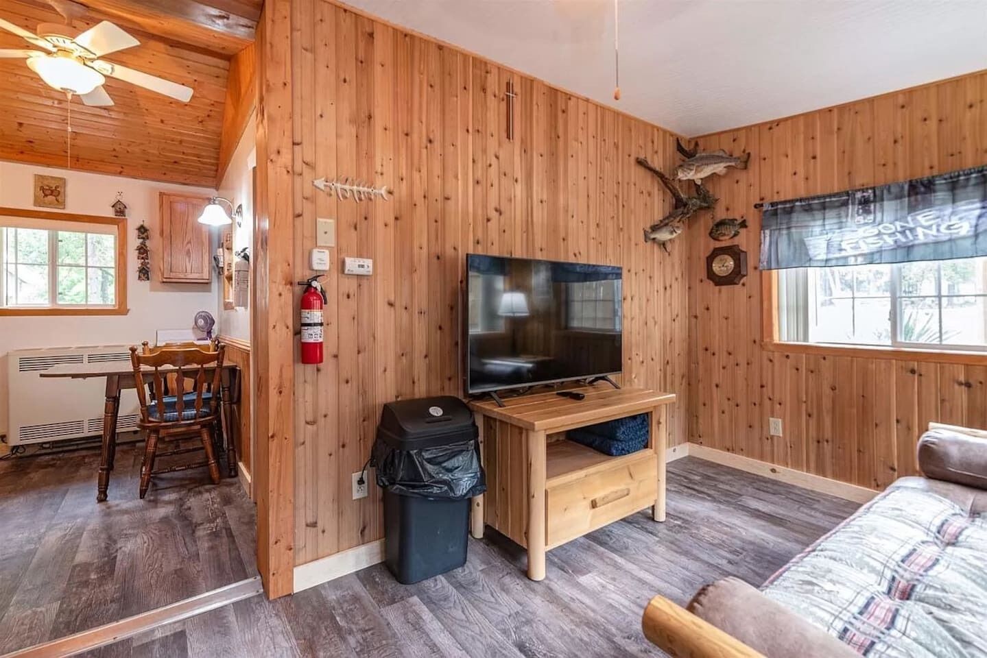 A room with wooden walls, a television on a wooden stand, a fire extinguisher, a trash bin, and a window with a view.