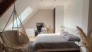 Un dormitorio moderno con techo inclinado, una silla colgante, una cama con ropa de cama gris y un escritorio pequeño con silla.