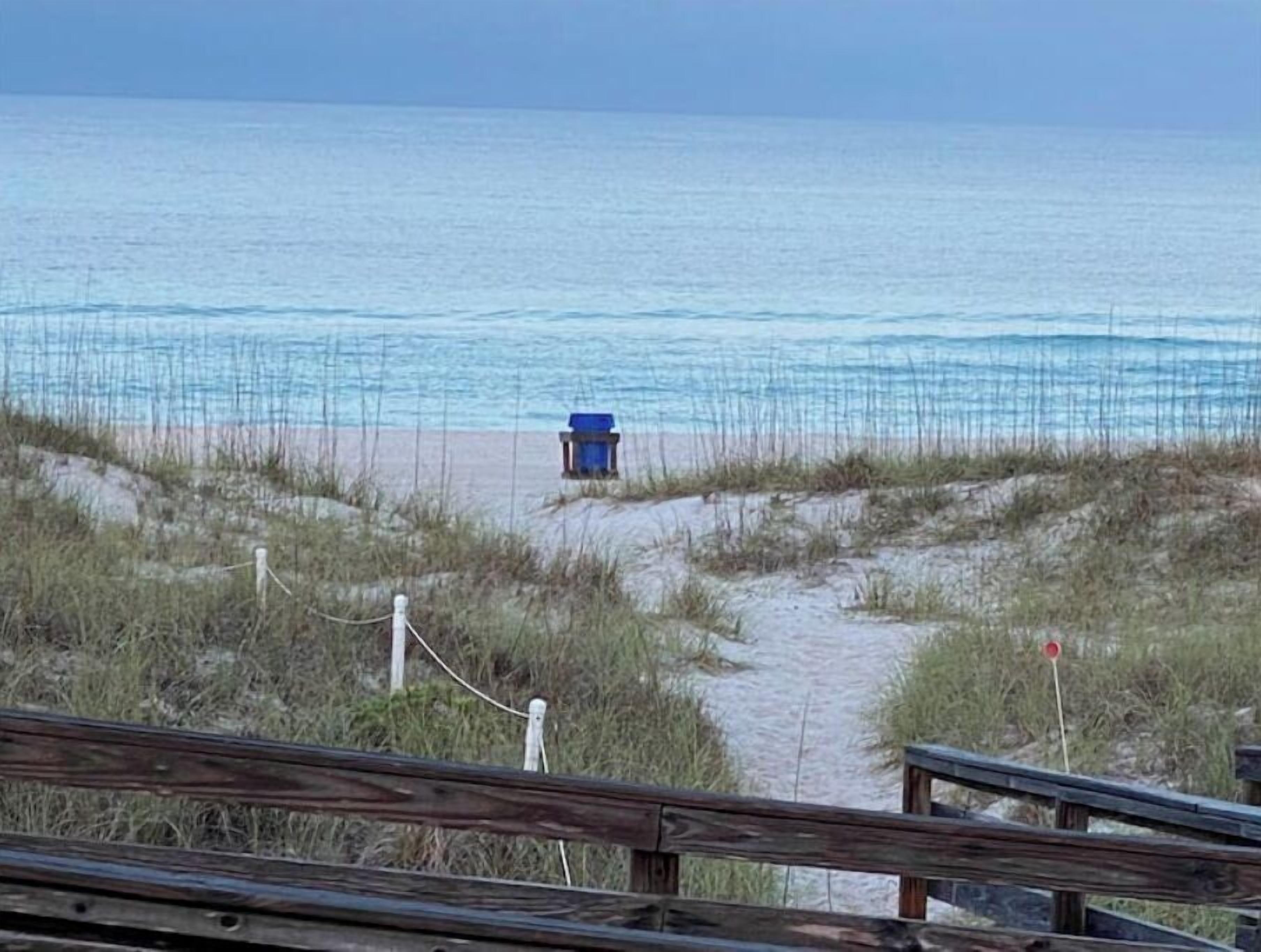 A beach path leading to the ocean with a blue trash bin and a wooden fence in the foreground.