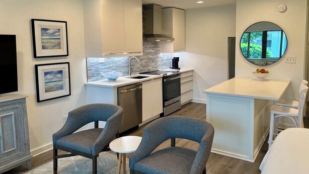 A modern kitchen with white cabinets, a built-in stove, and a round mirror above the sink. There are two gray chairs and a small white table. - Newly remodeled, heart of Seagrove Beach, 100 yards to beach! (Santa Rosa Beach)