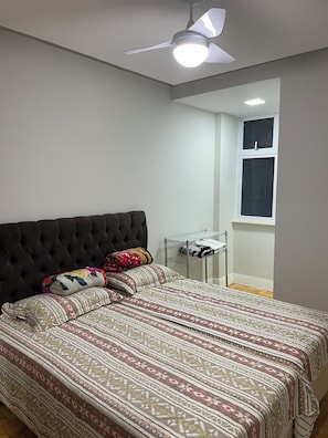 Quarto com cabeceira estofada, edredom estampado, ventoinha no teto e janela com persianas.