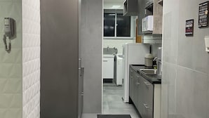Una cocina estrecha con paredes de azulejos blancos, una encimera negra y un microondas. Hay un tapete negro en el suelo que conduce a una puerta.