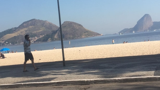Escena de playa con una persona caminando sobre la arena, una farola y montañas al fondo.