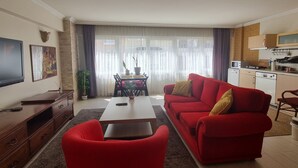 Una sala de estar con un sofá rojo, una mesa de centro y un televisor. Se observa un comedor con una mesa y sillas. La cocina es visible, con electrodomésticos y armarios.