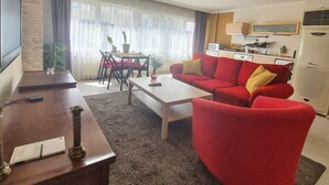 Sala de estar con un sofá rojo, una mesa de centro, una mesa de comedor con sillas, un área de cocina con electrodomésticos y un amplio ventanal con cortinas.