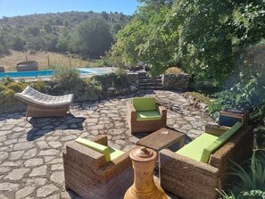 Un espacio al aire libre con mobiliario de mimbre y suelo de piedra, con vistas a una piscina y abundante vegetación.