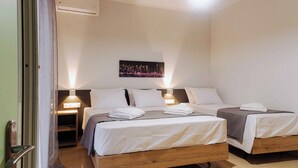 A hotel room with two beds, a nightstand with a lamp, an air conditioning unit, and a cityscape painting on the wall.