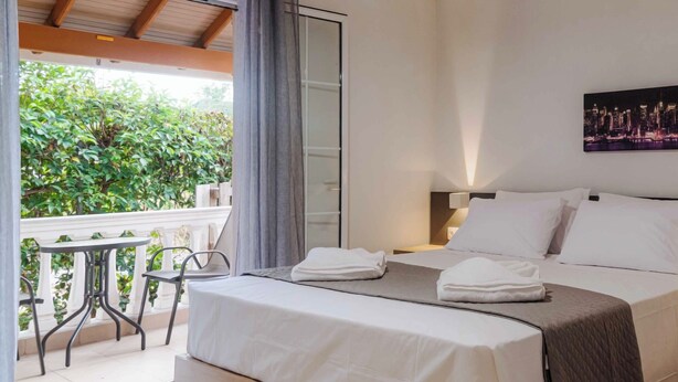 A hotel room with a bed, a balcony with a table and chairs, an air conditioning unit, and a painting on the wall.
