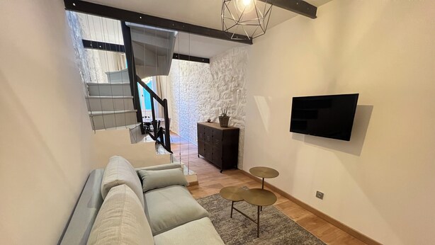 A modern living room with a sectional sofa, a staircase, a flat-screen TV mounted on the wall, and a small round table. - Maisonnette Centre Historique de Nîmes Tout Équipé ! (Nîmes)