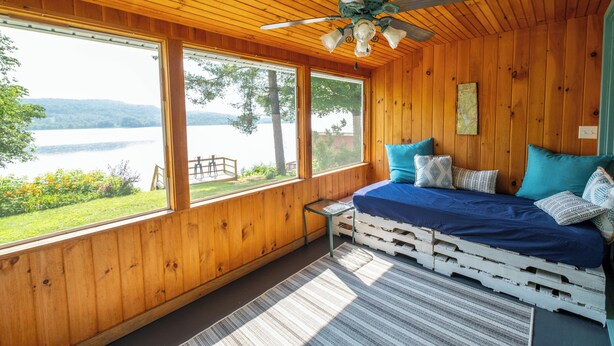 Una cabaña de madera con una cama hecha de palets, ventanas amplias y un ventilador de techo.