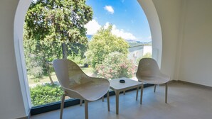 Une pièce avec deux fauteuils en osier et une petite table, de grandes fenêtres en arc offrant une vue sur les arbres et un bâtiment.