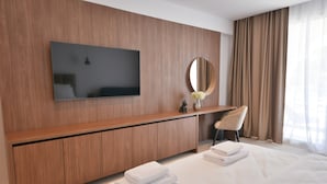 Une chambre d’hôtel moderne équipée d’un téléviseur à écran plat fixé au mur, d’une console en bois, d’un miroir rond, d’un bureau avec une chaise et de serviettes soigneusement pliées sur le lit.