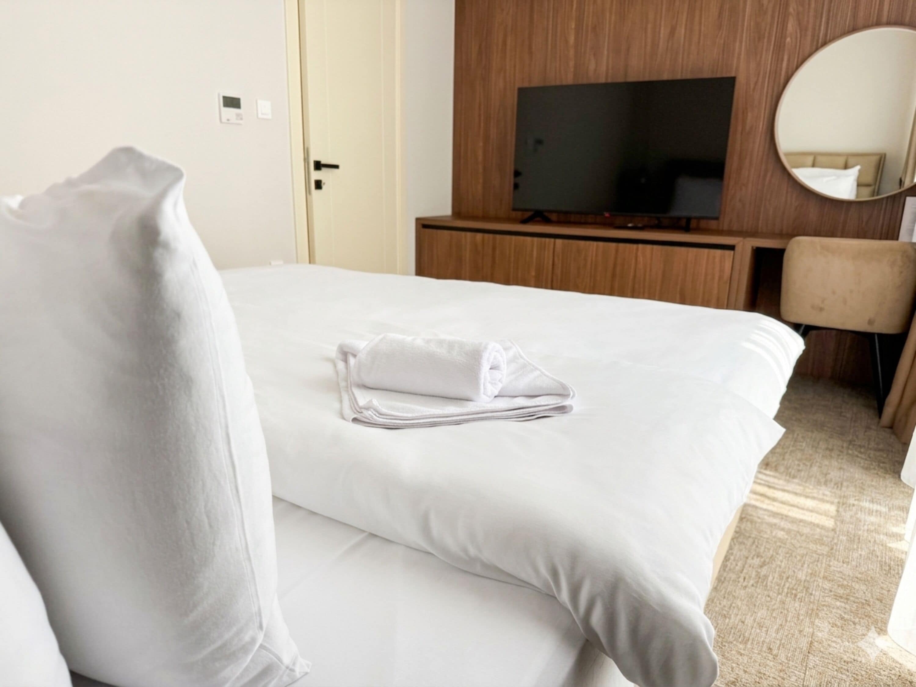 A hotel room with a neatly made bed, a television on a wooden stand, a chair, and a round mirror on the wall.