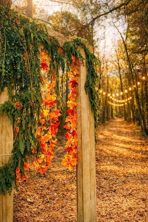 Un arco de madera adornado con hojas otoñales y follaje, rodeado por un sendero forestal con luces colgantes.