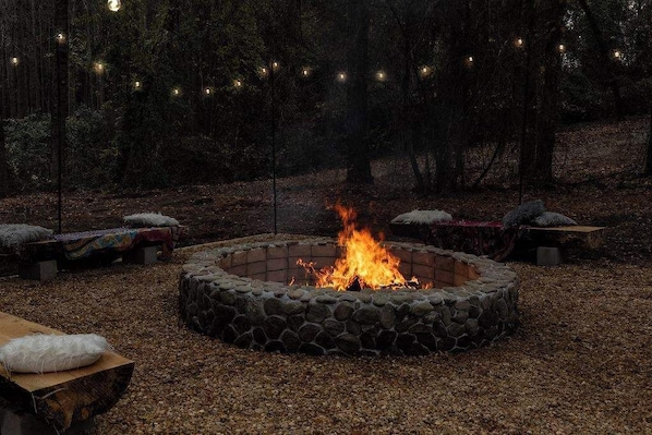 Un hogar de piedra rodeado de bancos y cojines en un área boscosa con luces colgantes.