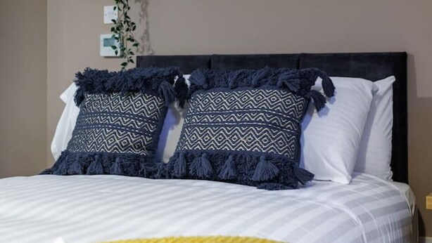 A neatly made bed with a white and yellow blanket, two patterned pillows, and a dark headboard. Above the bed, there is a wall-mounted sign with the word "hello".