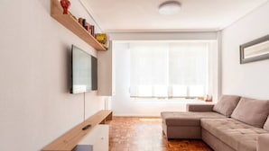 A modern living room with a sofa, a flat-screen TV, wooden flooring, and a large window. - Apartamento Canalejas (Santander)