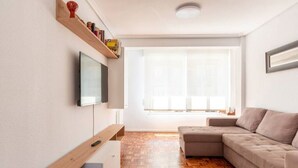 A modern living room with a wooden floor, a beige sofa, a TV mounted on the wall, and a window with sheer curtains. - Apartamento Canalejas (Santander)