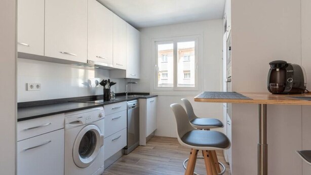 A compact kitchen with white cabinets, a washing machine, a coffee maker, and a small dining area with two chairs. - Apartamento Canalejas (Santander)