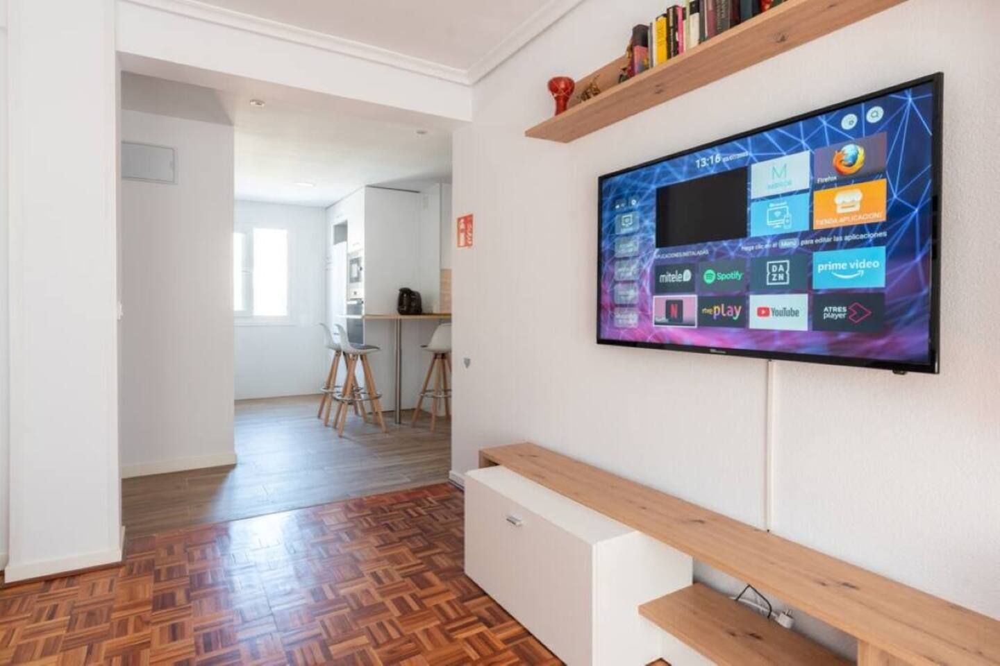 Una sala moderna con un televisor de pantalla plana montado en la pared que muestra varios servicios de streaming, un banco de madera y un suelo con estampado tipo espiga.
