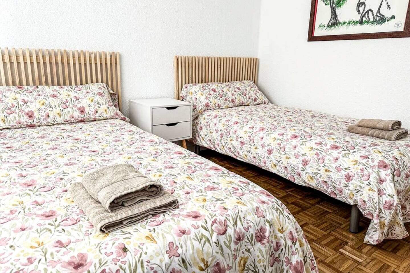 Una habitación con dos camas, cada una con ropa de cama floral y cabeceros de madera, una mesita de noche pequeña y una obra de arte colgada en la pared.