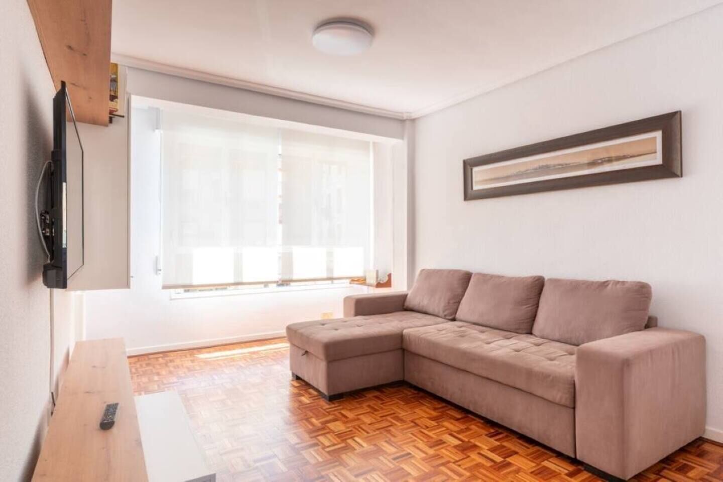 A living room with a sectional sofa, a flat-screen TV mounted on the wall, and a large window with blinds.