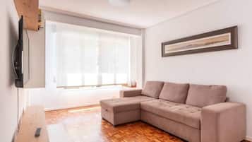 A living room with a sectional sofa, a flat-screen TV mounted on the wall, and a large window with blinds.