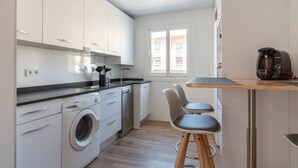 A compact kitchen with white cabinets, a washing machine, a coffee maker, and a small dining area with stools. - Apartamento Canalejas (Santander)