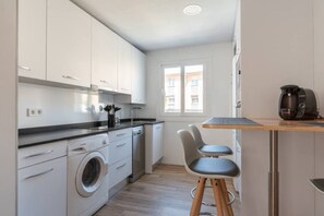 A compact kitchen with white cabinets, a washing machine, a coffee maker, and a small dining area with stools. - Apartamento Canalejas (Santander)