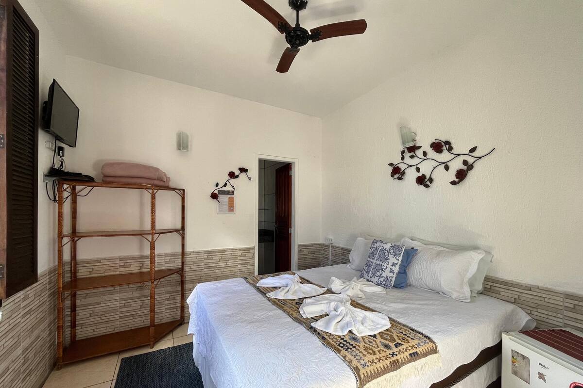 A hotel room with a bed, a ceiling fan, a TV, a wooden shelf, and a decorative wall piece.