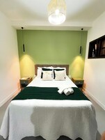 Una cama bien hecha con colcha verde y sábanas blancas, dos mesitas de noche con lámparas, y un candelabro colgante en el techo.