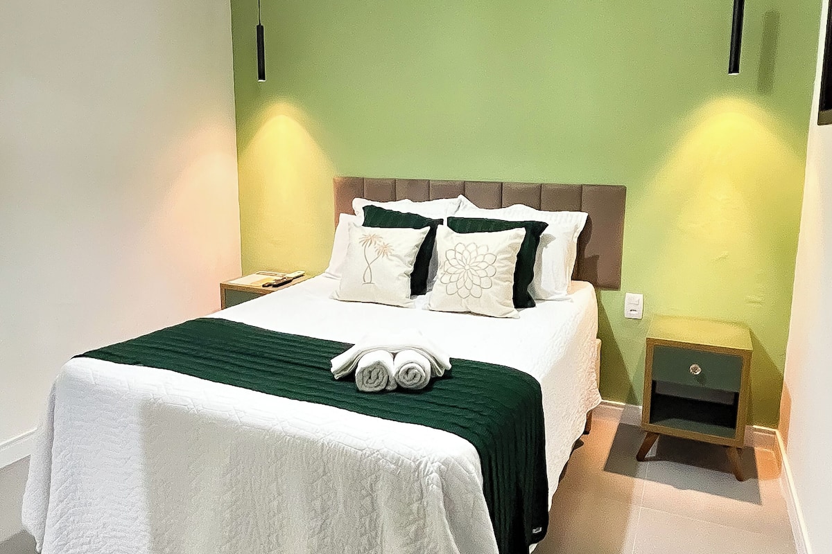 A neatly made bed with a green and white bedspread, two pillows, and rolled towels. There is a small bedside table with a lamp, and a green wall with a hanging light fixture.