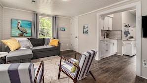 A living room with a gray sofa, striped armchair, and a painting of a pelican. A kitchen with white cabinets and a tiled backsplash is visible through an open doorway.