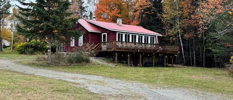Une cabane rustique dotée d’une terrasse en bois et d’un toit rouge, entourée d’arbres aux feuilles d’automne.