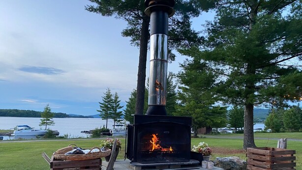 A wood-burning stove with a fire, surrounded by wooden crates and a bench, set on a deck with a view of a lake and boats.