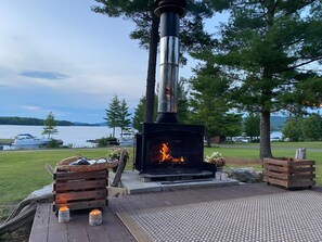 Una estufa de leña con fuego, rodeada de cajas de madera y un banco, ubicada en una terraza con vistas a un lago y barcos.