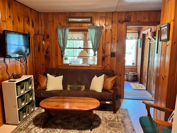 Una habitación acogedora de estilo cabaña, con interior de madera, un sofá con cojines, un televisor montado en la pared y una ventana con cortinas.