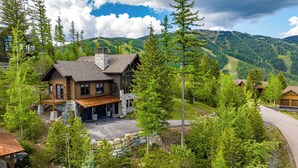 Una grande casa a due piani in legno, con fondamenta in pietra e un garage, immersa nel verde e circondata da montagne.