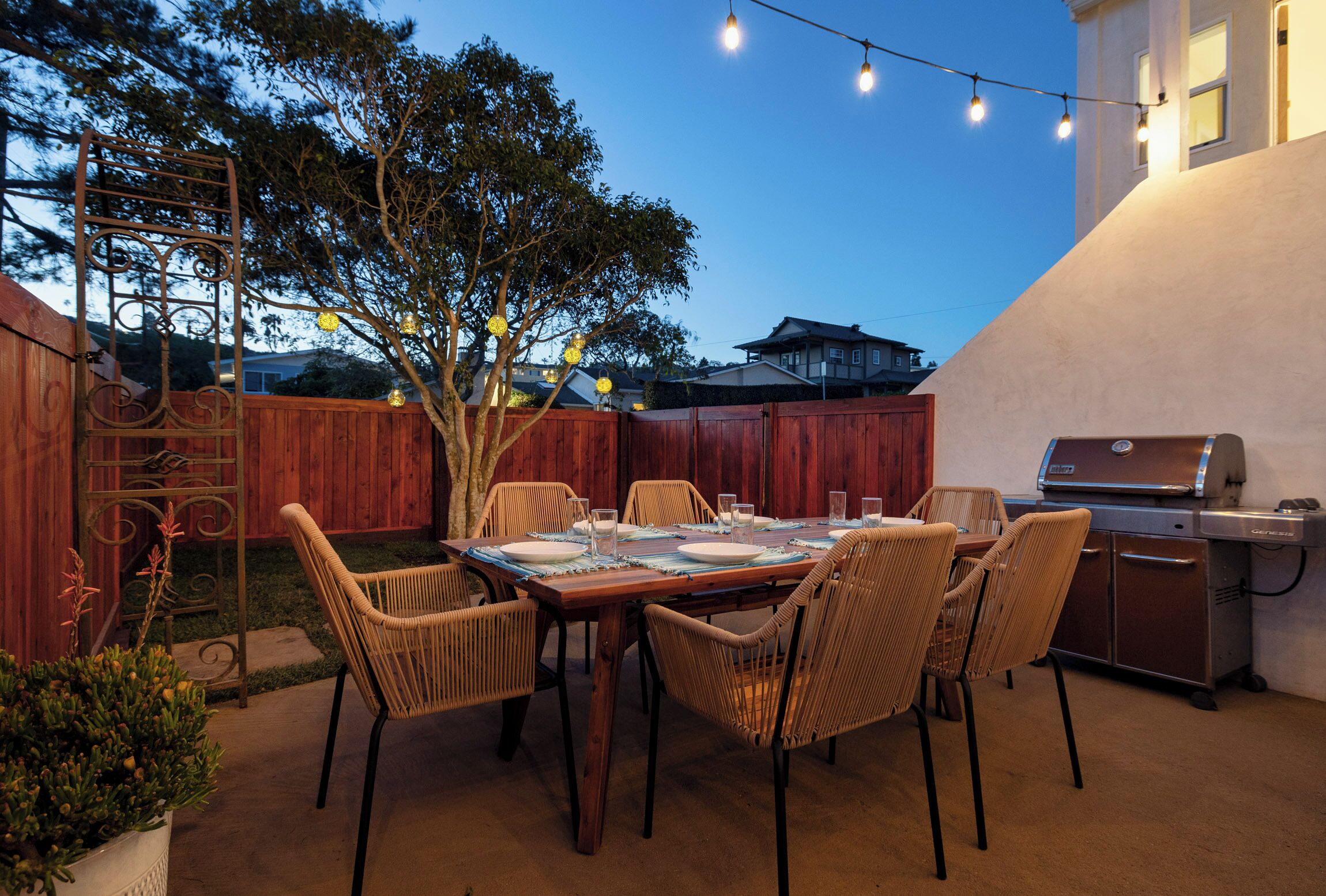 Une terrasse extérieure avec une table en bois prévue pour quatre personnes, entourée de chaises en osier, d’un barbecue et de guirlandes lumineuses.