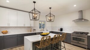 Een moderne keuken met grijze kastjes, een wit eiland, twee barkrukken, een gasfornuis en hangende lampen.