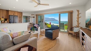 Un moderno salón con un gran ventanal con vistas panorámicas al océano, un ventilador de techo y una cocina con armarios de madera.