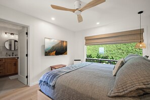 Ein Schlafzimmer mit Bett, Deckenventilator, Fenster mit Blick auf Grünflächen und einem Gemälde an der Wand.