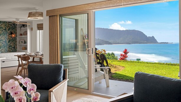 Una sala de estar moderna con un gran ventanal corredera que da a un balcón con vistas al océano y a las montañas.
